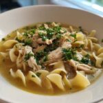 A close-up of a white bowl filled with rich broth, egg noodles, shredded chicken, and fresh parsley, showcasing the amazing 1-pot chicken noodle soup.