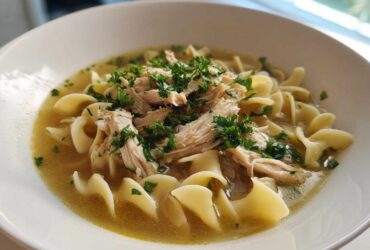 A close-up of a white bowl filled with rich broth, egg noodles, shredded chicken, and fresh parsley, showcasing the amazing 1-pot chicken noodle soup.