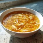 A close-up of a steaming white bowl filled with golden broth and delicate ribbons of egg drop soup.