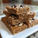 A stack of five homemade, chewy protein bars topped with oats and chocolate chips on a white plate.
