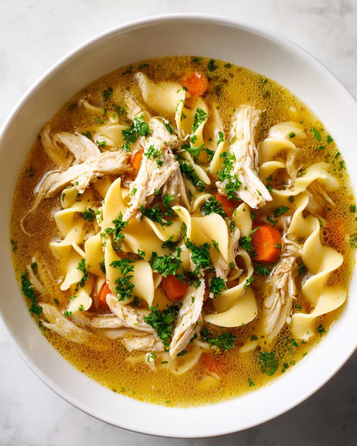 A close-up overhead view of a white bowl filled with rich broth, egg noodles, shredded chicken, and carrots in amazing 1-pot chicken noodle soup.