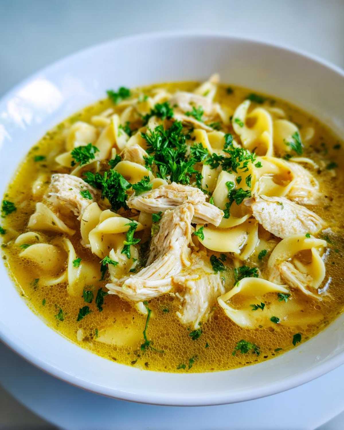 A close-up view of a white bowl filled with rich broth, egg noodles, shredded chicken, and fresh parsley, showcasing the amazing chicken noodle soup.