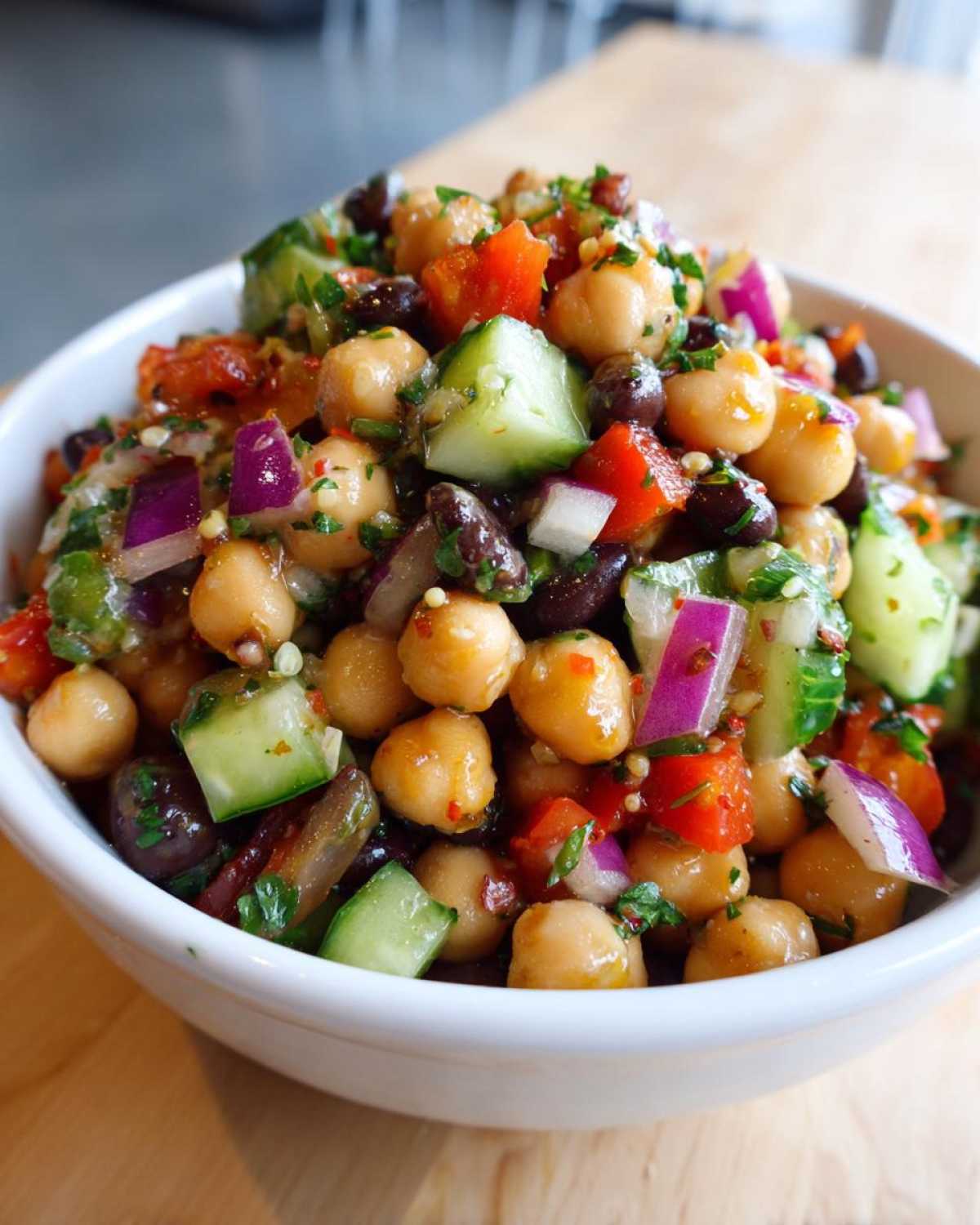 Close-up of a vibrant, fresh bean salad featuring chickpeas, black beans, cucumber, red onion, and tomatoes in a white bowl.