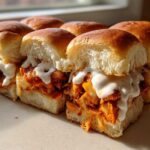 A close-up of several baked buffalo chicken sliders filled with shredded chicken, melted cheese, and drizzled with ranch or blue cheese dressing.