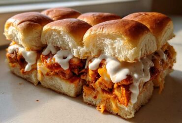 A close-up of several baked buffalo chicken sliders filled with shredded chicken, melted cheese, and drizzled with ranch or blue cheese dressing.