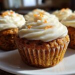 A close-up of several moist carrot cake muffins topped with thick cream cheese frosting and orange zest.
