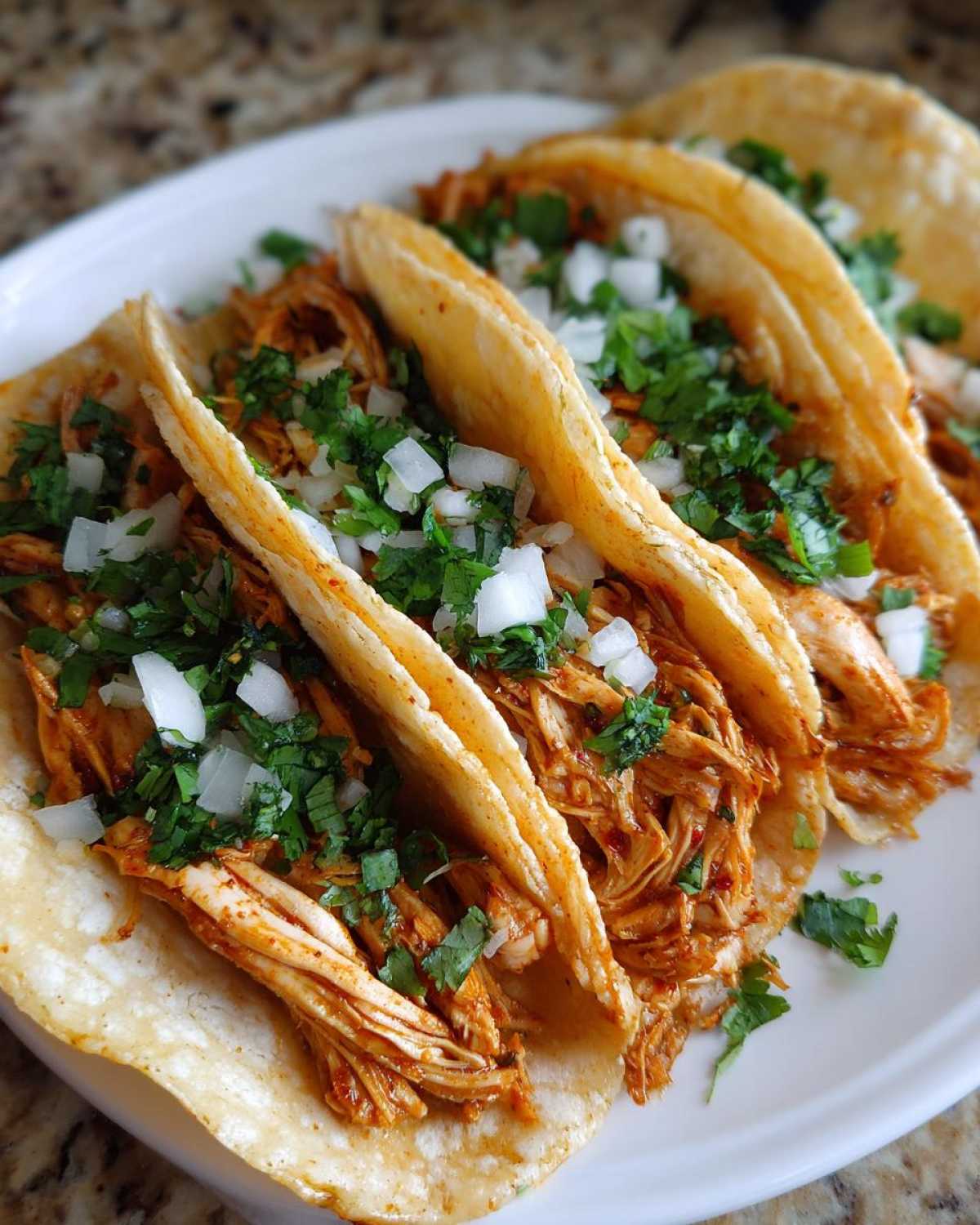 Close-up of four soft corn tortillas filled with shredded chicken tacos, topped with diced white onion and cilantro.