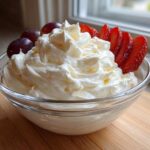 A glass bowl filled with creamy, white fruit dip, garnished with sliced strawberries and grapes.