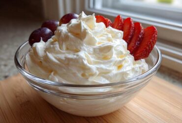 A glass bowl filled with creamy, white fruit dip, garnished with sliced strawberries and grapes.