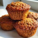 A close-up stack of four golden brown, moist honey bran muffins sitting on a white plate near a window.