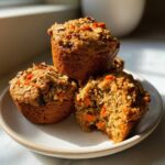 Three delicious morning glory muffins stacked on a plate, one with a bite taken out showing carrots and nuts.