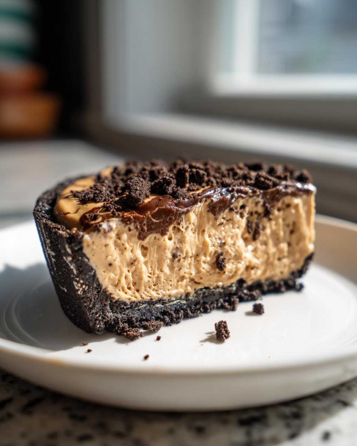 A close-up of a slice of no-bake peanut butter pie with a dark cookie crust, topped with chocolate drizzle and crumbs.