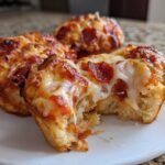 Close-up of three freshly baked pizza muffins, one broken open showing gooey cheese and pepperoni topping.