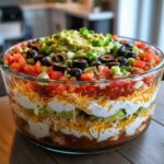 Close-up of a towering Seven layer dip in a clear glass bowl, topped with guacamole, olives, and tomatoes.