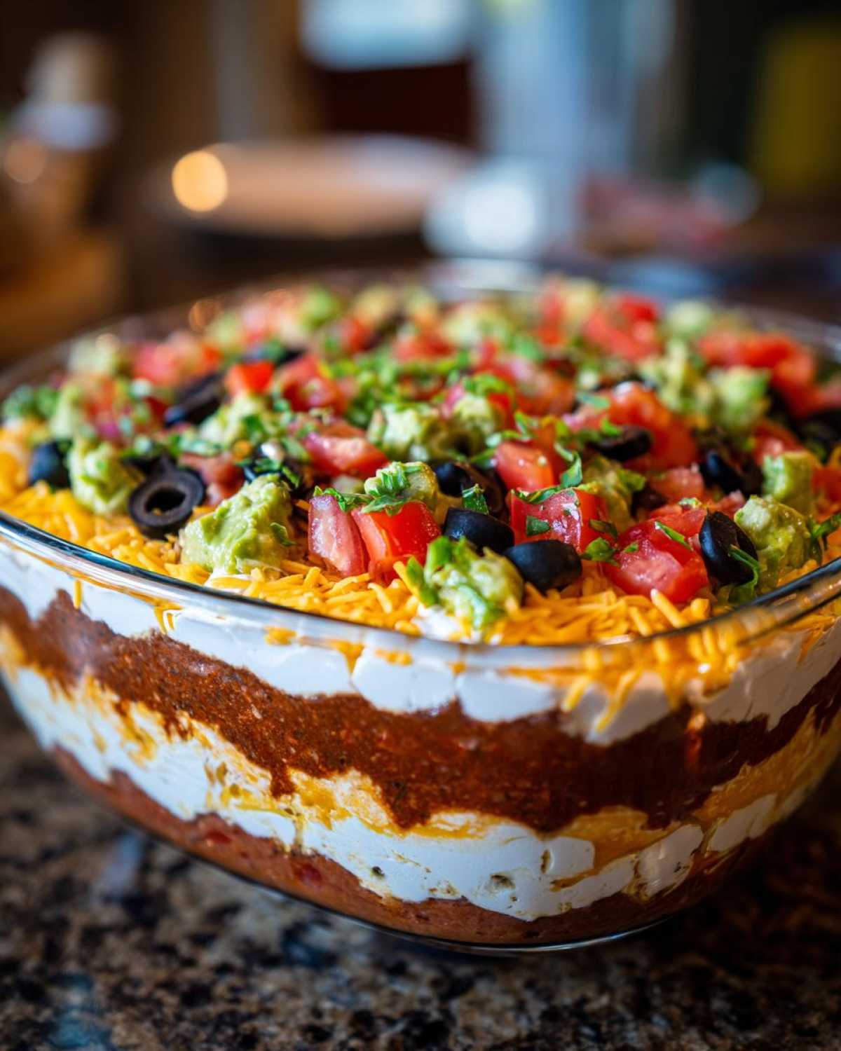 Close-up view of a clear glass dish filled with Amazing Seven layer dip, showing distinct layers and toppings like guacamole, tomatoes, and olives.