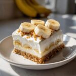 A square slice of layered banana cream pie recipe with a graham cracker crust, cream filling, and fresh banana slices on top.