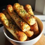 Close-up of freshly baked, golden garlic breadsticks seasoned with parsley, piled high in a white bowl.