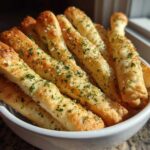 A close-up of freshly baked, golden breadsticks seasoned with garlic, cheese, and parsley, served upright in a white bowl.