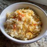 A close-up view of a white bowl filled with creamy, fluffy cheesy rice topped with shredded cheddar cheese.