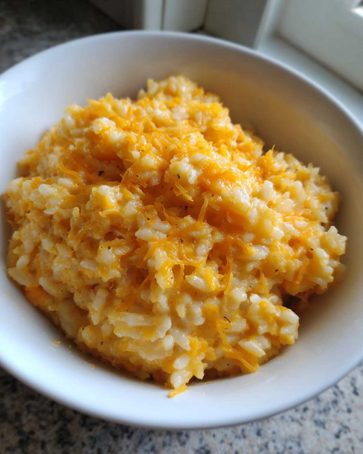 Close-up of a white bowl filled with creamy cheesy rice topped generously with bright orange shredded cheddar cheese.