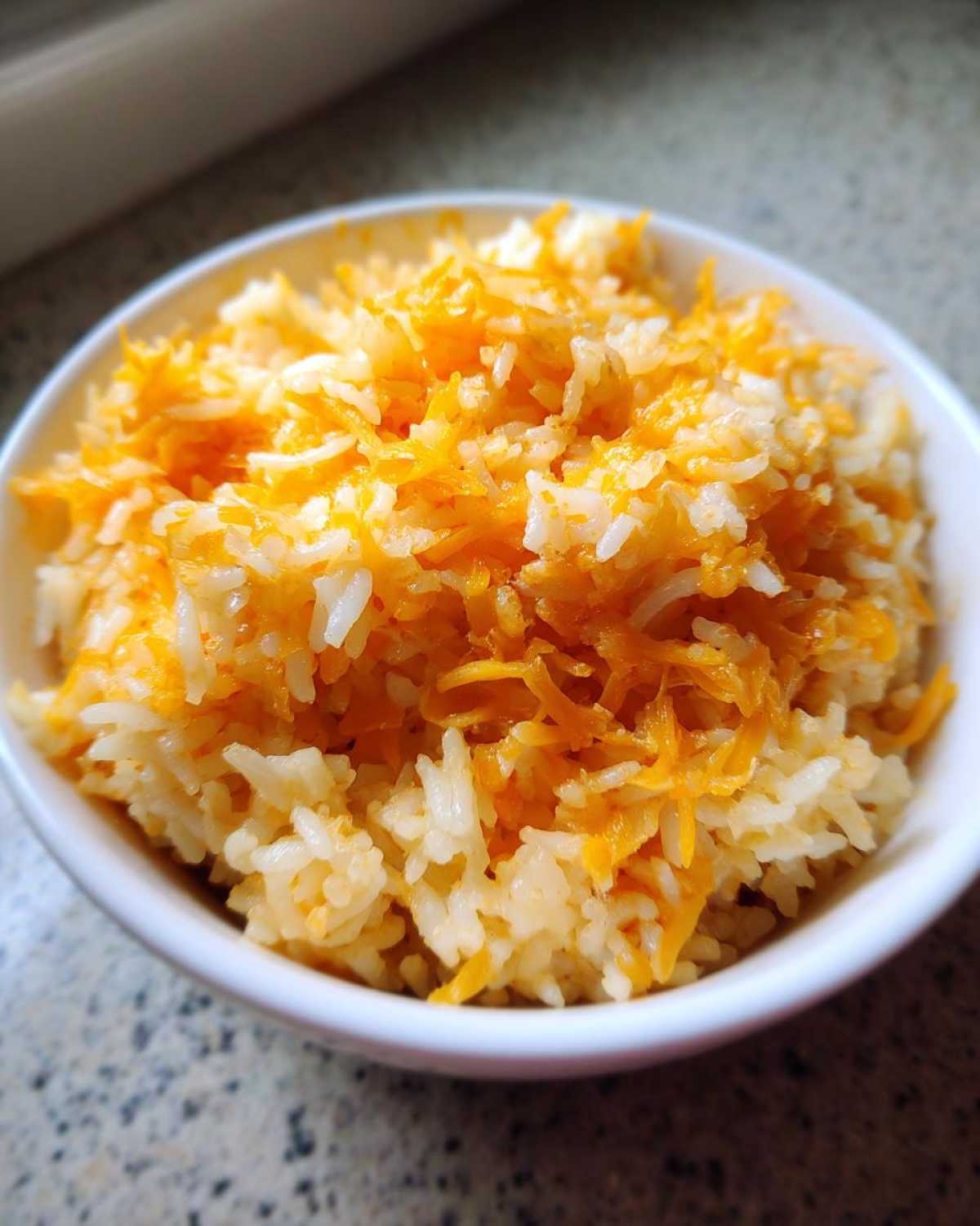 Close-up of a white bowl filled with fluffy white rice topped generously with bright orange shredded cheddar cheese, making perfect cheesy rice.
