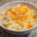 A white bowl filled with creamy, cheesy rice topped with bright orange shredded cheese and pepper.