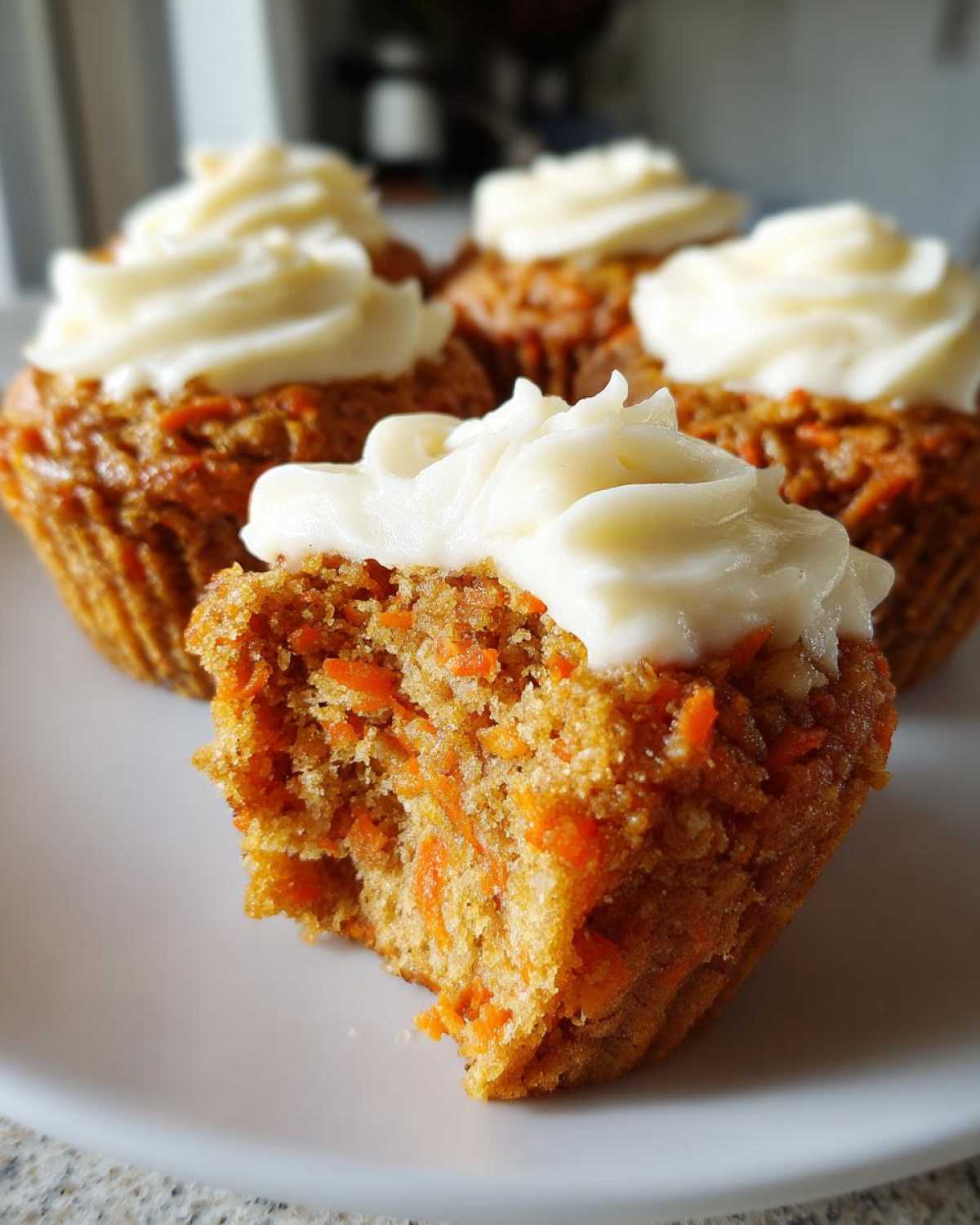 A bitten carrot cake muffin showing moist texture and shredded carrots, topped with cream cheese frosting, with more carrot cake muffins in the background.
