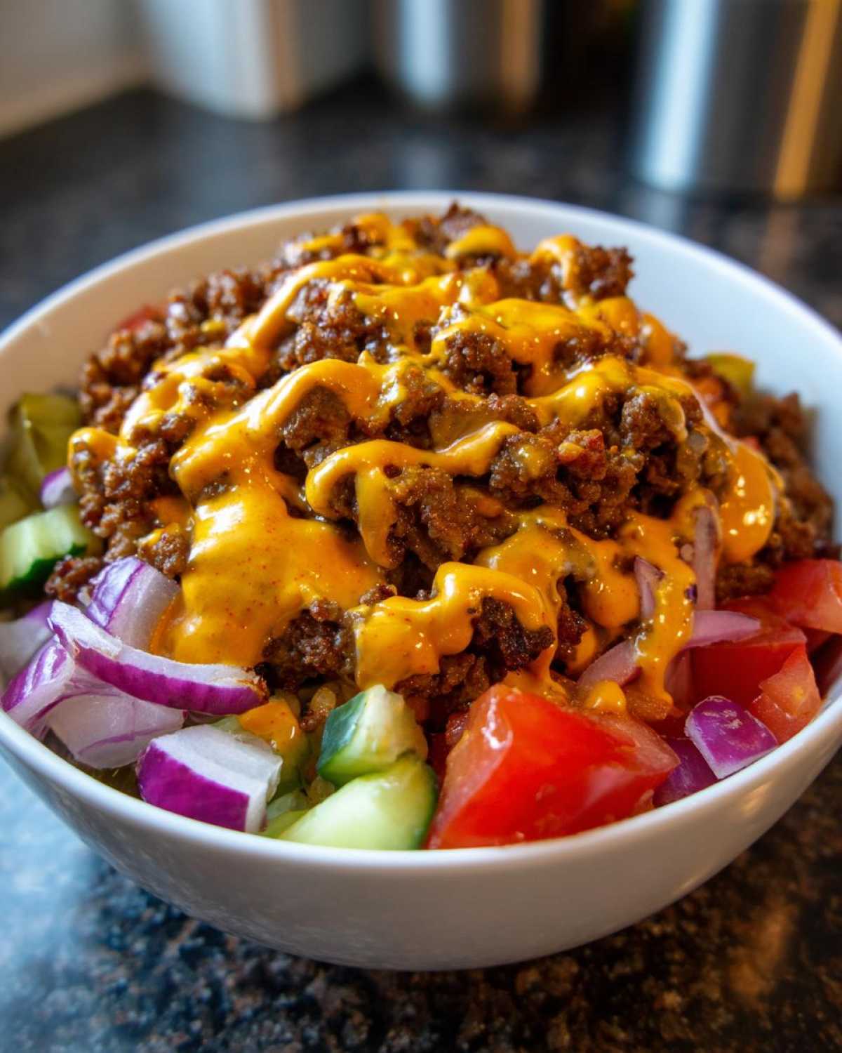 A close-up view of a white bowl filled with a cheeseburger bowl, topped with seasoned ground beef, melted cheese sauce, tomatoes, cucumbers, and red onion.