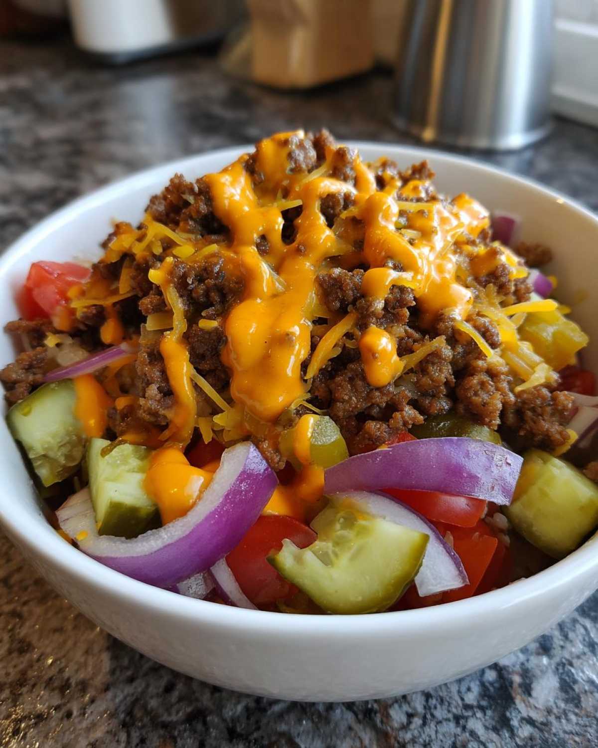A white bowl filled with a cheeseburger bowl featuring seasoned ground beef, melted cheese sauce, pickles, red onion, and tomatoes.