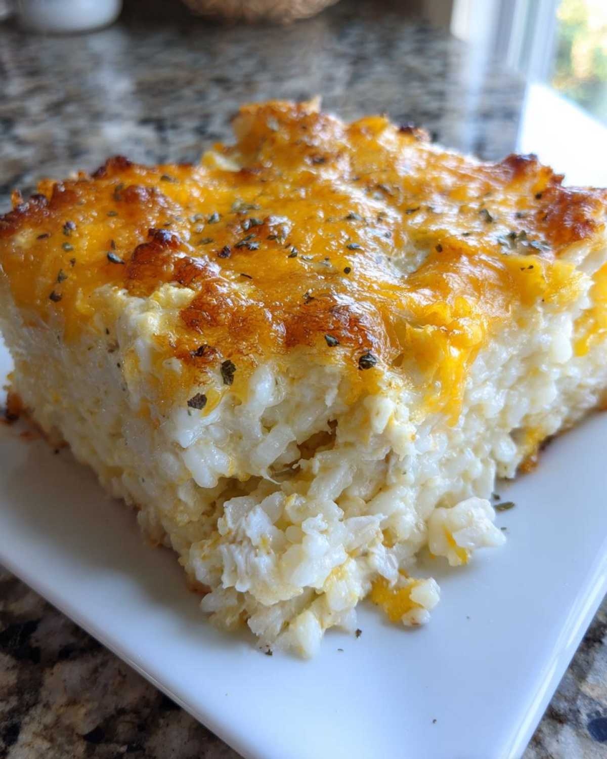 A close-up of a square serving of cheesy chicken and rice casserole with a golden-brown melted cheddar topping.