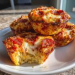 A stack of golden-brown pizza muffins topped with melted cheese and pepperoni, one has a bite taken out showing the cheesy interior.