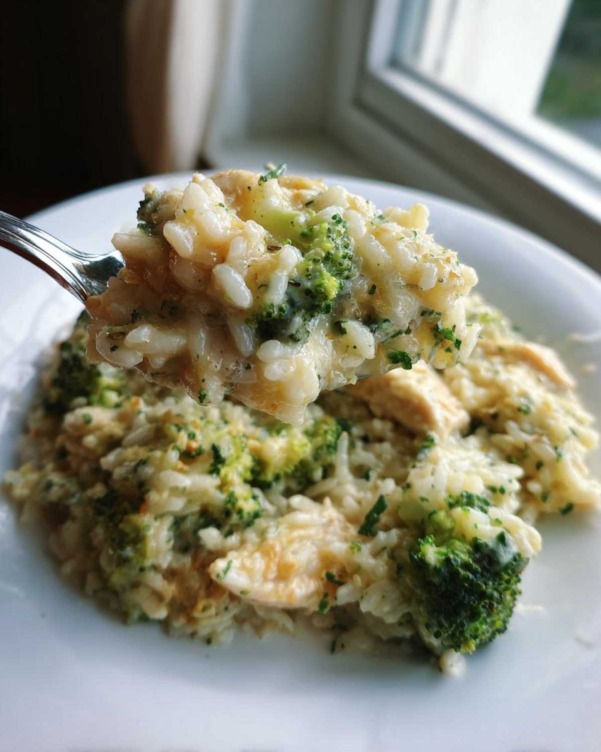 A spoonful of creamy chicken broccoli rice casserole being lifted above a white plate.