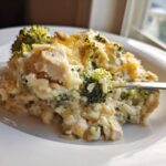 A spoonful of creamy chicken broccoli rice casserole topped with melted cheddar cheese being lifted from a white plate.