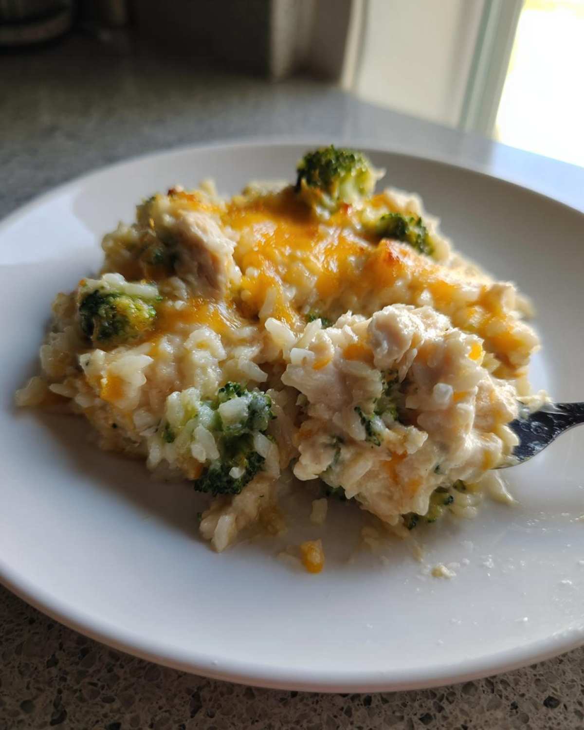 Close-up of a serving of creamy chicken broccoli rice casserole topped with melted cheddar cheese, being lifted by a fork.