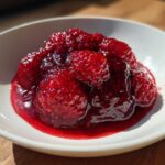Close-up of chunky, vibrant red raspberry jam with whole berries in a white bowl.