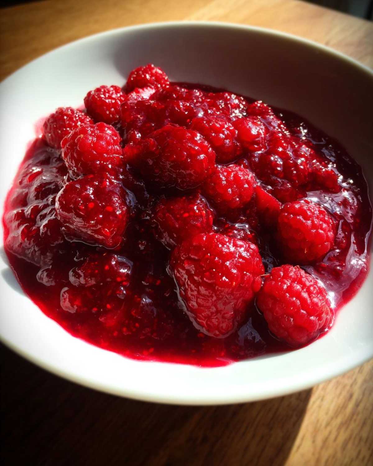 Close-up of vibrant, whole raspberries suspended in thick, glossy homemade raspberry jam in a white bowl.
