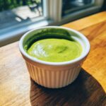 A close-up of vibrant green cilantro lime sauce served in a small white fluted ramekin on a wooden surface.
