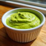 A close-up of thick, bright green cilantro lime sauce served in a small white ramekin on a wooden surface.