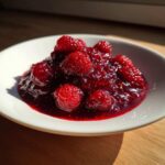 A small white bowl filled with glistening, deep red raspberry jam topped with whole, fresh raspberries.