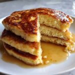 Close-up of a stack of three thick coconut flour pancakes, cut to show the texture, drizzled with maple syrup.