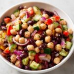 Close-up view of a vibrant, colorful bean salad featuring chickpeas, kidney beans, cucumber, red onion, and bell peppers.