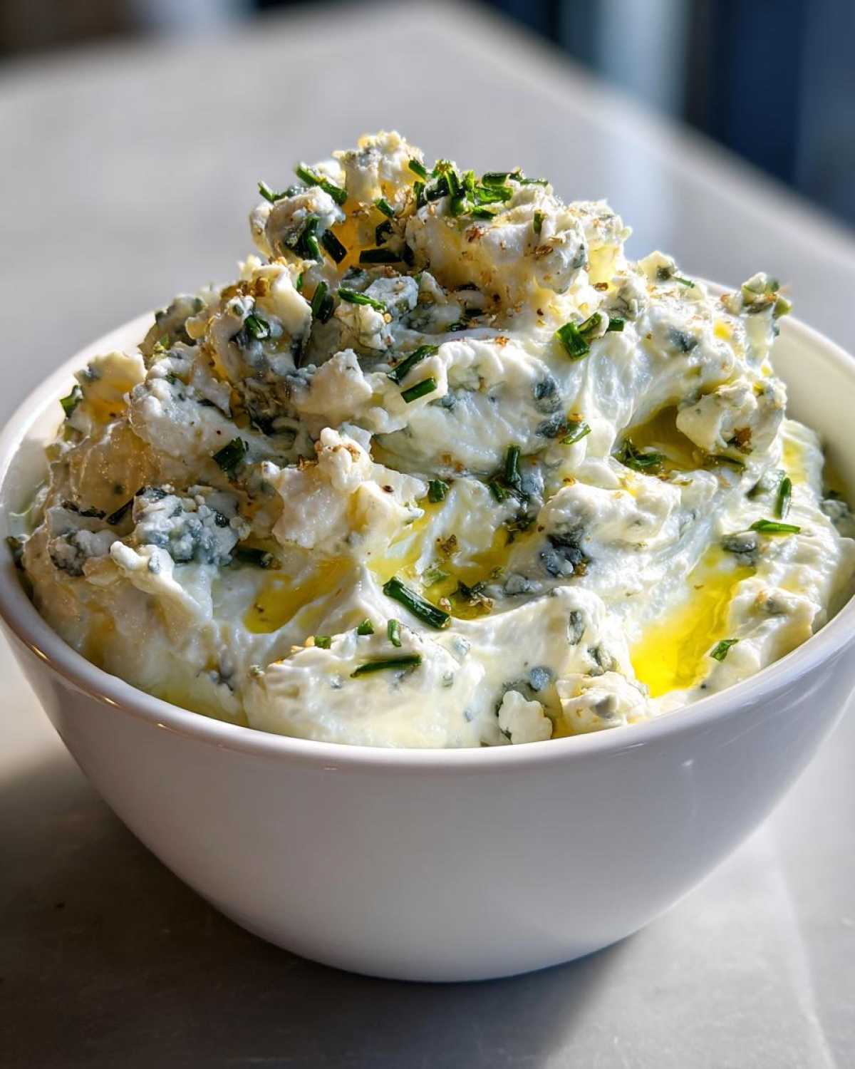 Close-up of a white bowl filled with a creamy, chunky blue cheese mixture, drizzled with olive oil and topped with fresh chives for the dirty martini dip.