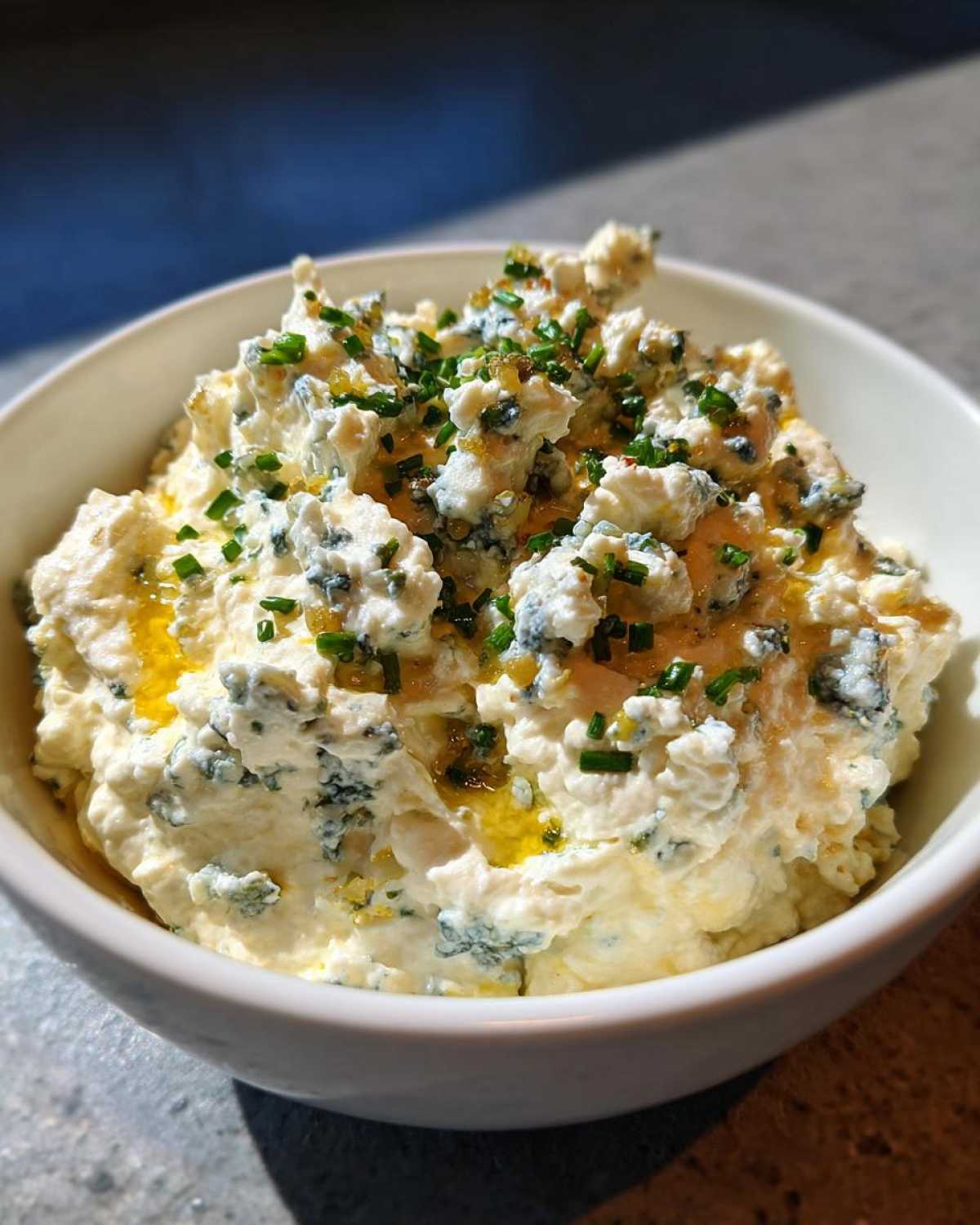 Close-up of a creamy, chunky dip featuring blue cheese crumbles, drizzled with oil and topped with chopped chives, likely the dirty martini dip.