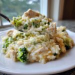 A spoonful of creamy chicken broccoli rice casserole lifted from a white plate, showing chunks of chicken and bright green broccoli.