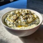 A white bowl filled with creamy dirty martini dip, topped generously with crumbled blue cheese and olive oil.