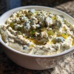 Close-up of a white bowl filled with creamy dirty martini dip topped with blue cheese crumbles and olive pieces.
