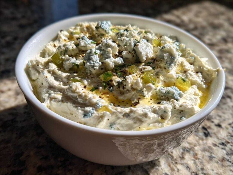 Close-up of a white bowl filled with creamy dirty martini dip topped with blue cheese crumbles and olive pieces.