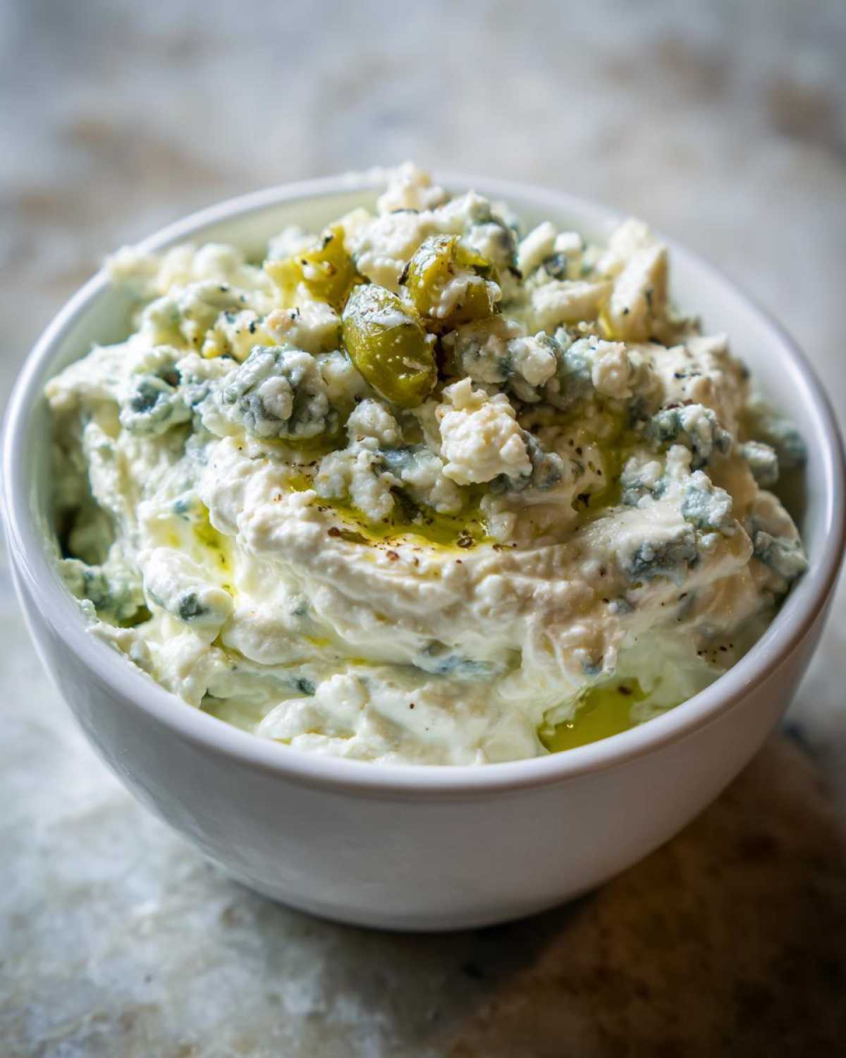 Close-up of a creamy dirty martini dip topped with blue cheese crumbles, green olives, and a drizzle of oil.