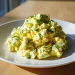 A generous serving of creamy, yellow egg salad recipe mixed with green onions and topped with fresh dill sprigs.