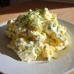 A generous serving of creamy egg salad recipe, mixed with green onions and topped with fresh dill, sits on a white plate.